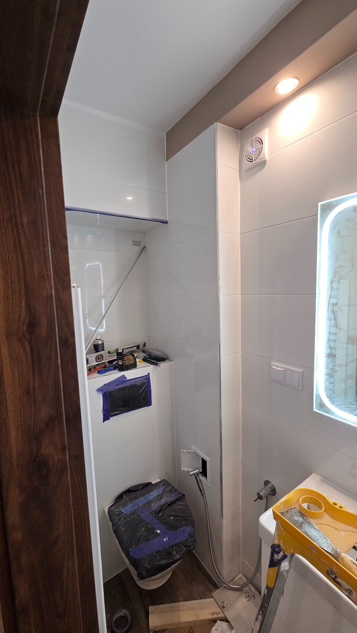 Biała łazienka w trakcie remontu: zabezpieczona toaleta, narzędzia na półce, świecące lustro. Widoczne płytki i wentylator. Częściowo widoczne drewniane obramowanie drzwi.