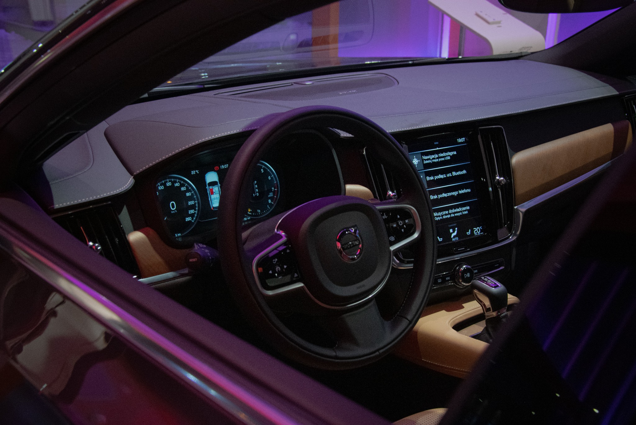 Wnętrze samochodu Volvo z widocznym kokpitem, kierownicą, ekranem nawigacji i beżową tapicerką, oświetlone fioletowym światłem.