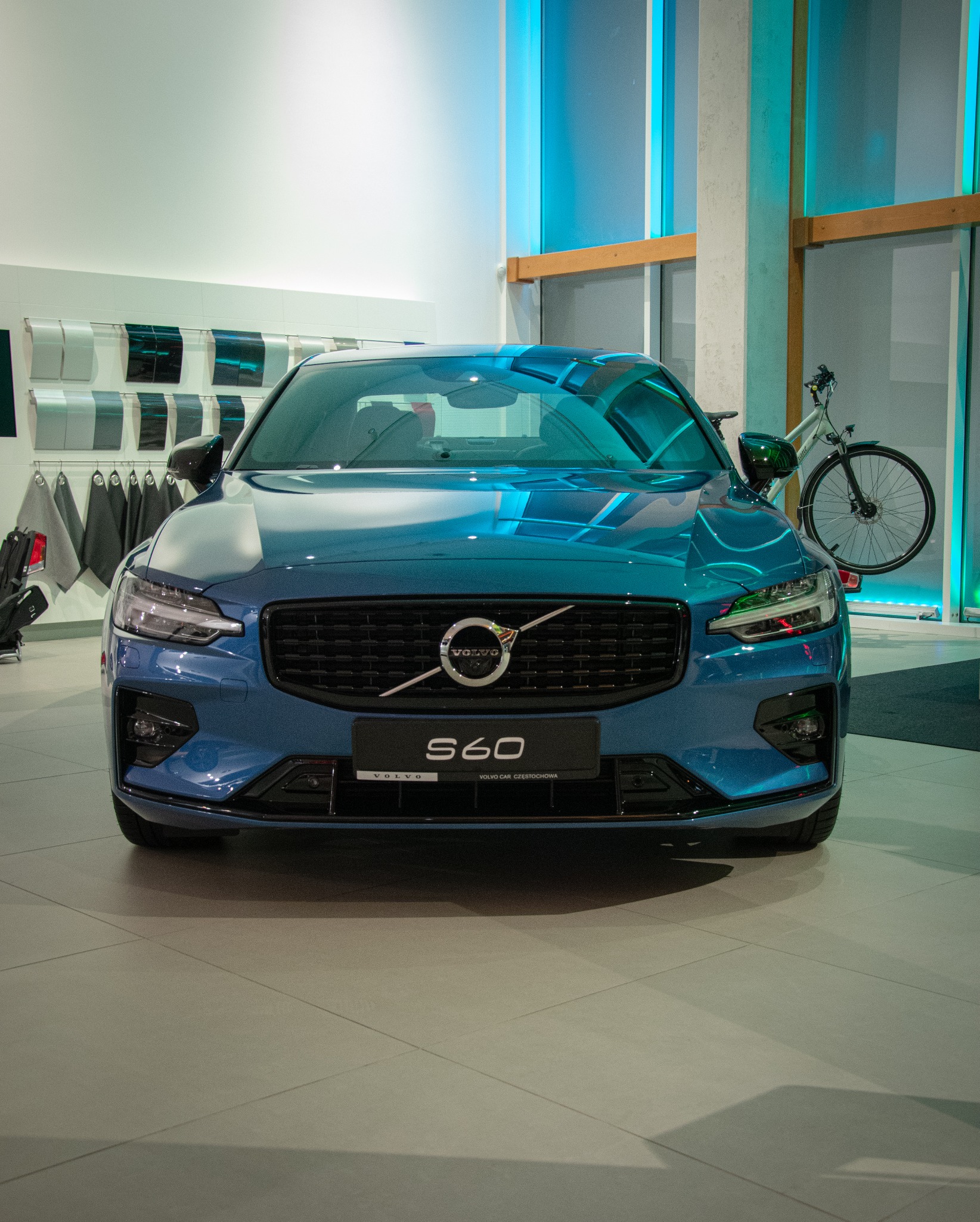 Niebieski samochód Volvo S60 sfotografowany z przodu w salonie samochodowym, z widocznymi próbkami kolorów lakieru i rowerem w tle.