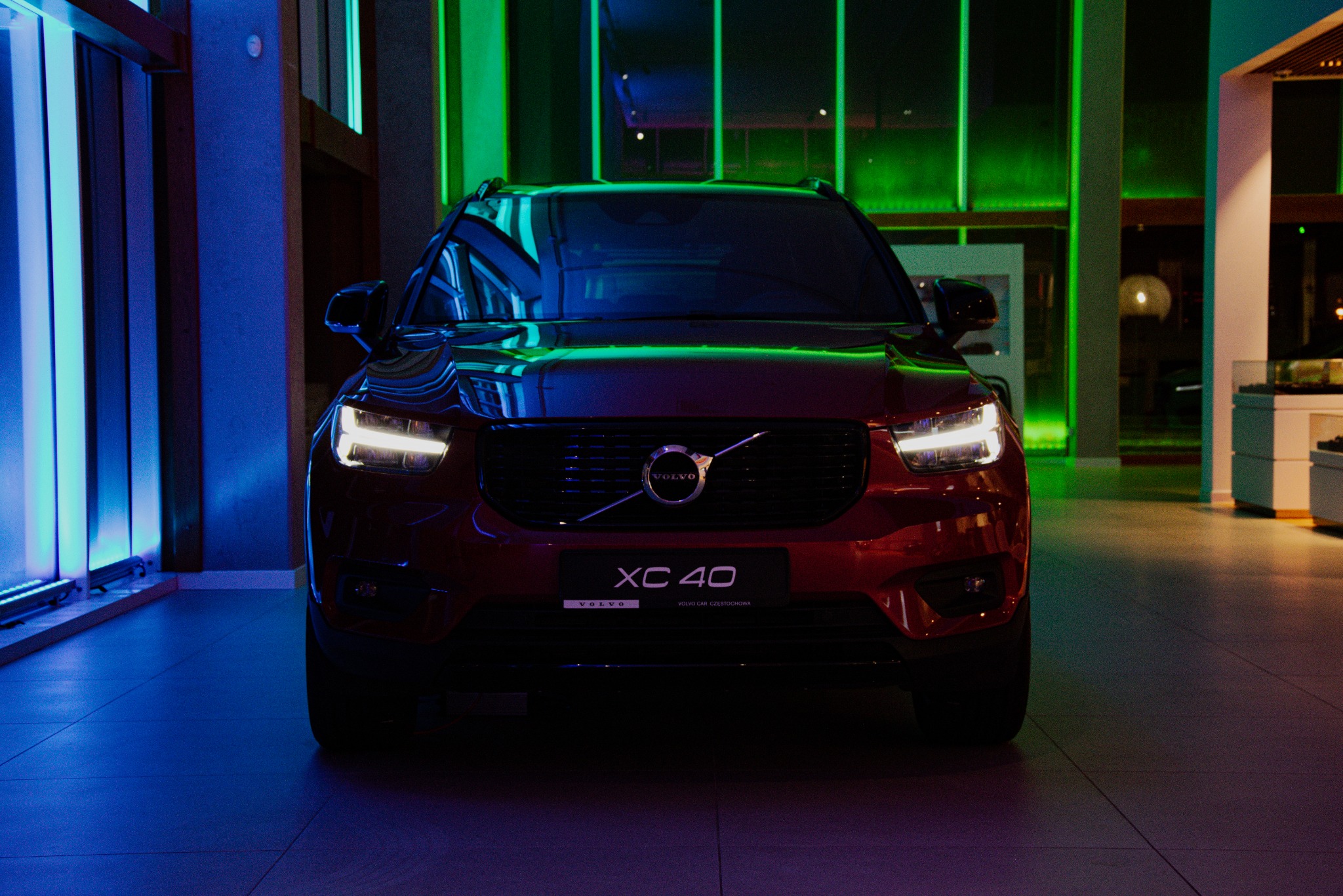 Czerwony samochód Volvo XC40 sfotografowany z przodu w salonie samochodowym, oświetlony niebieskim i zielonym światłem LED.