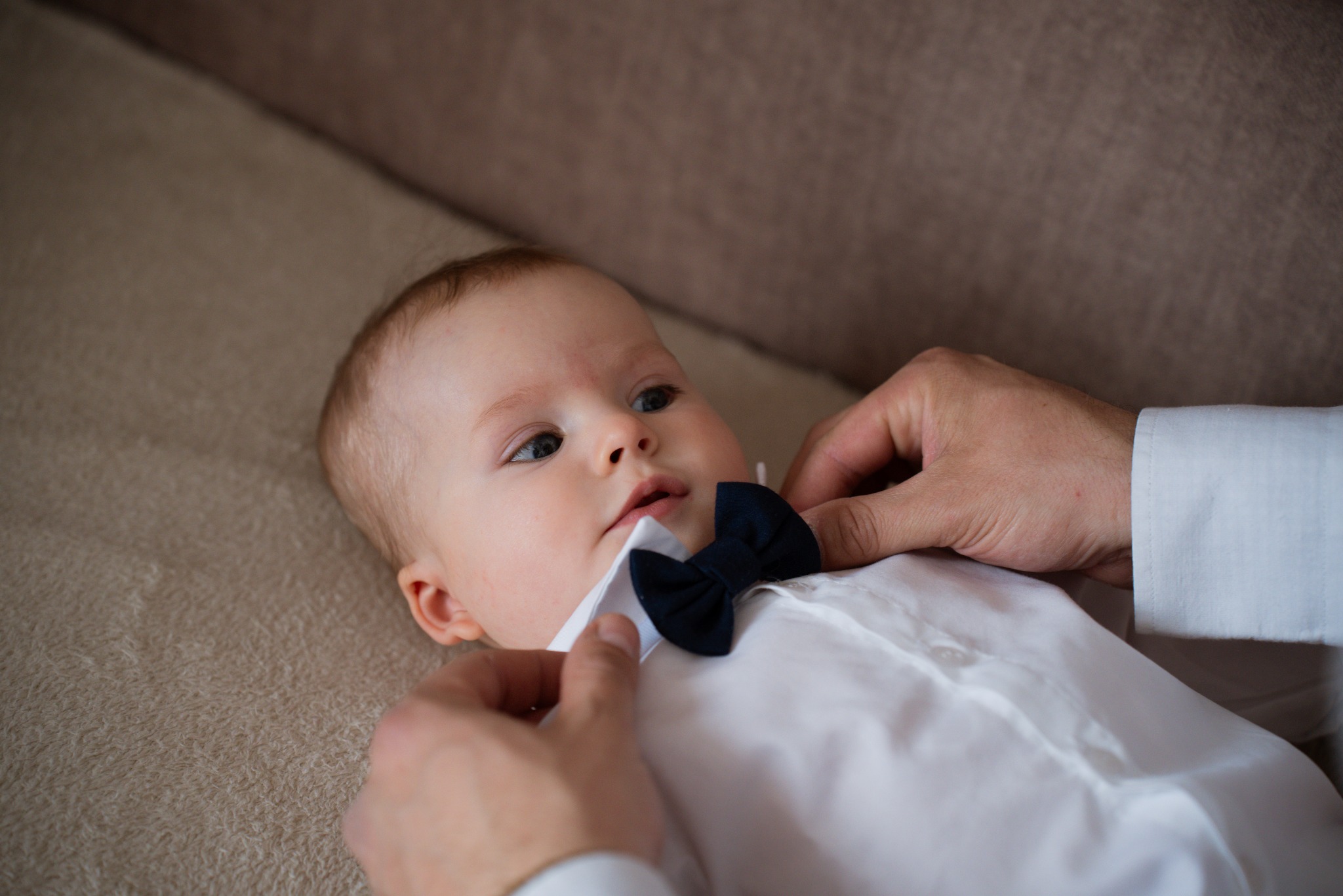 Uroczy niemowlak w białej koszuli i granatowej muszce, poprawianej przez dłonie dorosłego, leżący na beżowym tle. Delikatne światło podkreśla detale.
