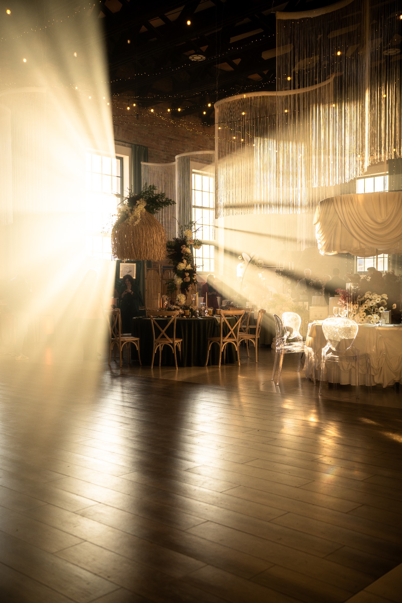 Uroczysta sala z dekoracjami kwiatowymi i światłem słonecznym wpadającym przez okna. Parkiet odbija blask, tworząc ciepłą atmosferę.