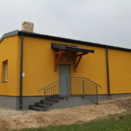 Żółty budynek z szarym cokołem, schodami i poręczą, zadaszeniem nad wejściem i dwoma kominami, otoczony zielenią i piaskiem.