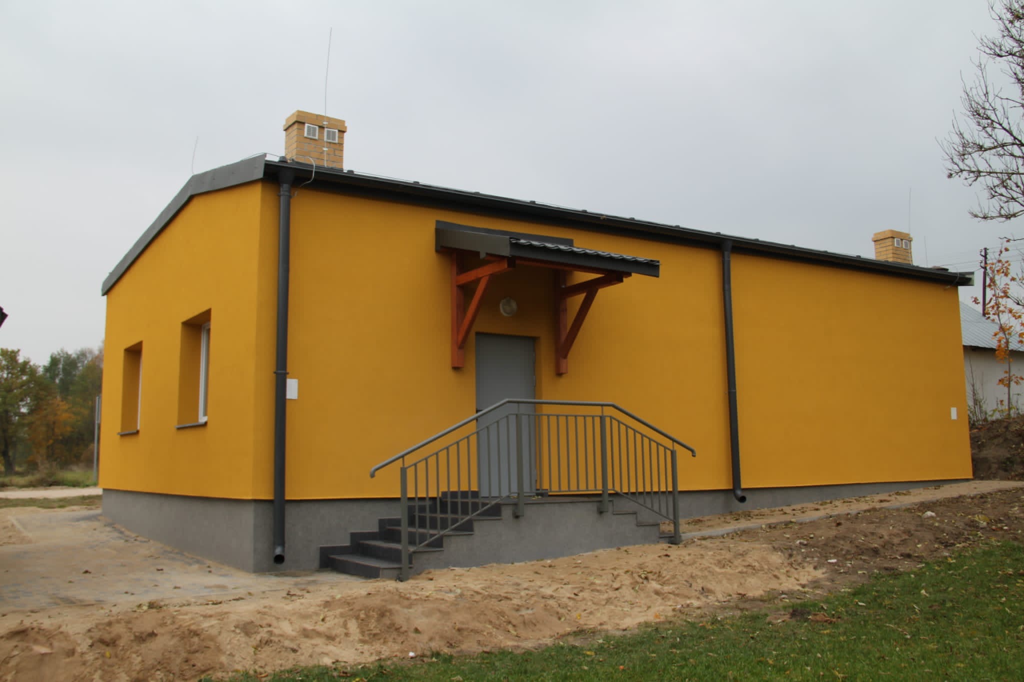 Żółty budynek z szarym cokołem, schodami i poręczą, zadaszeniem nad wejściem i dwoma kominami, otoczony zielenią i piaskiem.