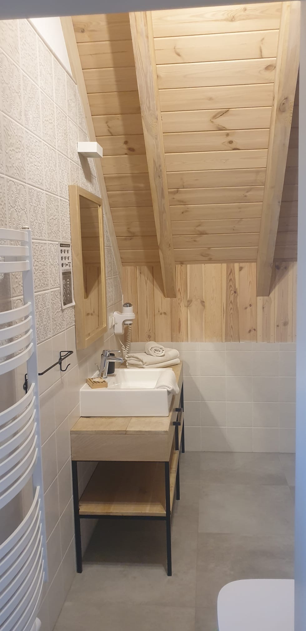 Jasna łazienka na poddaszu z drewnianym wykończeniem sufitu i ściany, białą umywalką na drewnianym stelażu z czarnymi nogami, grzejnikiem drabinkowym i dekoracyjnymi płytkami na ścianie.
