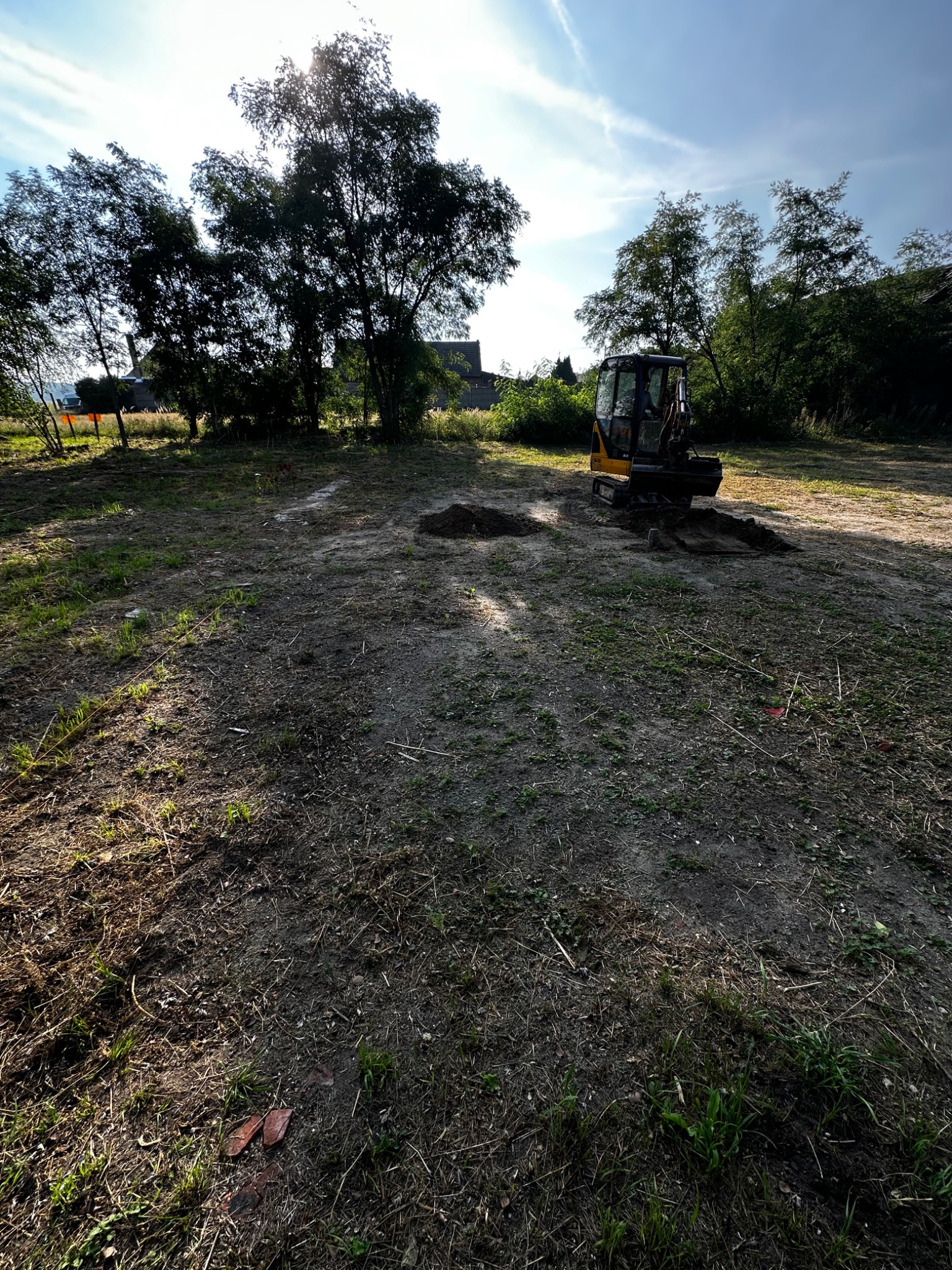 Żółto-czarna mini koparka na placu budowy, wykopane dołki, drzewa w tle, słoneczny dzień.