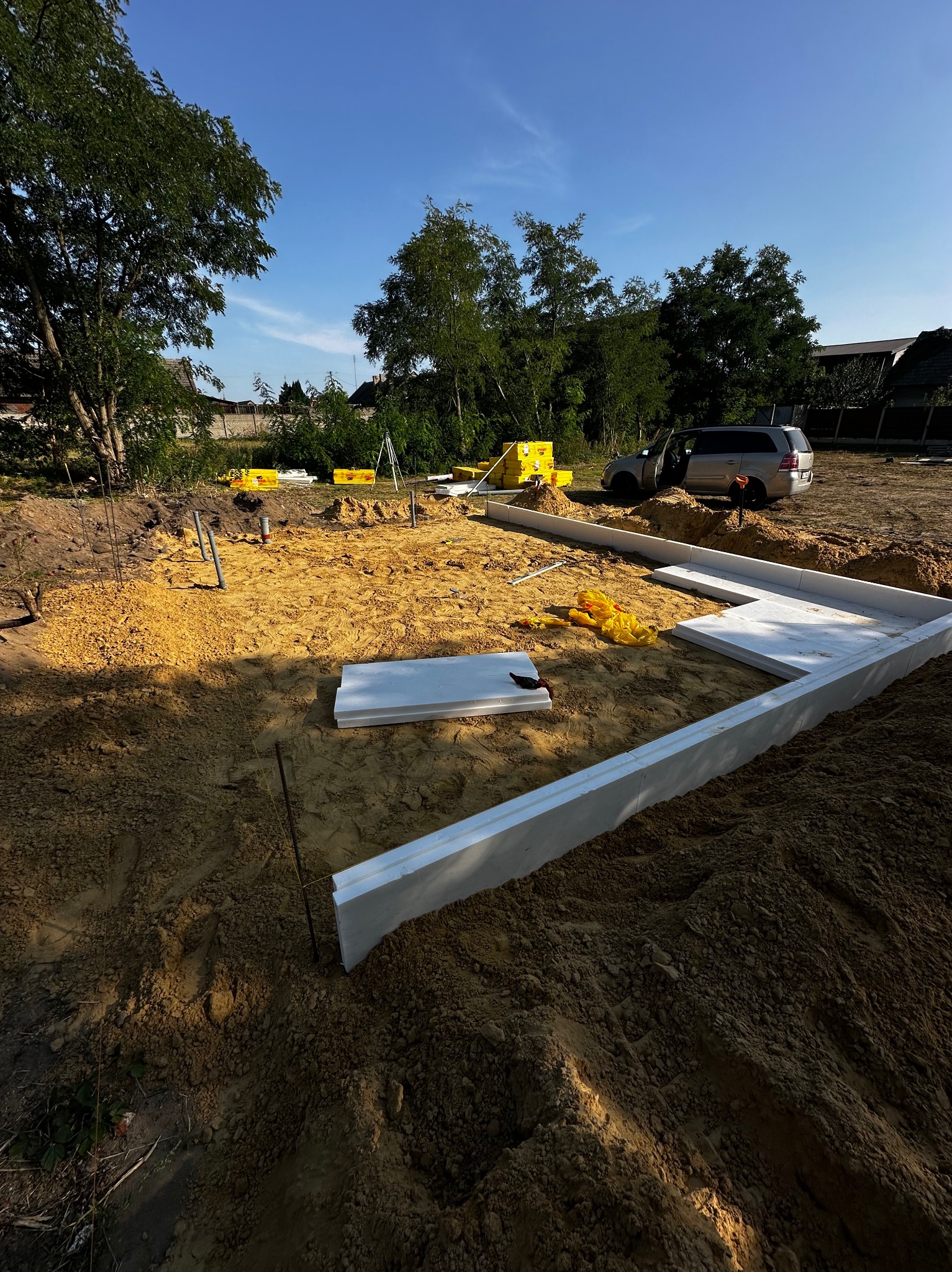 Przygotowany teren pod budowę z ułożonymi białymi panelami izolacyjnymi, widoczne wystające rury, samochód w tle, żółte worki i drzewa