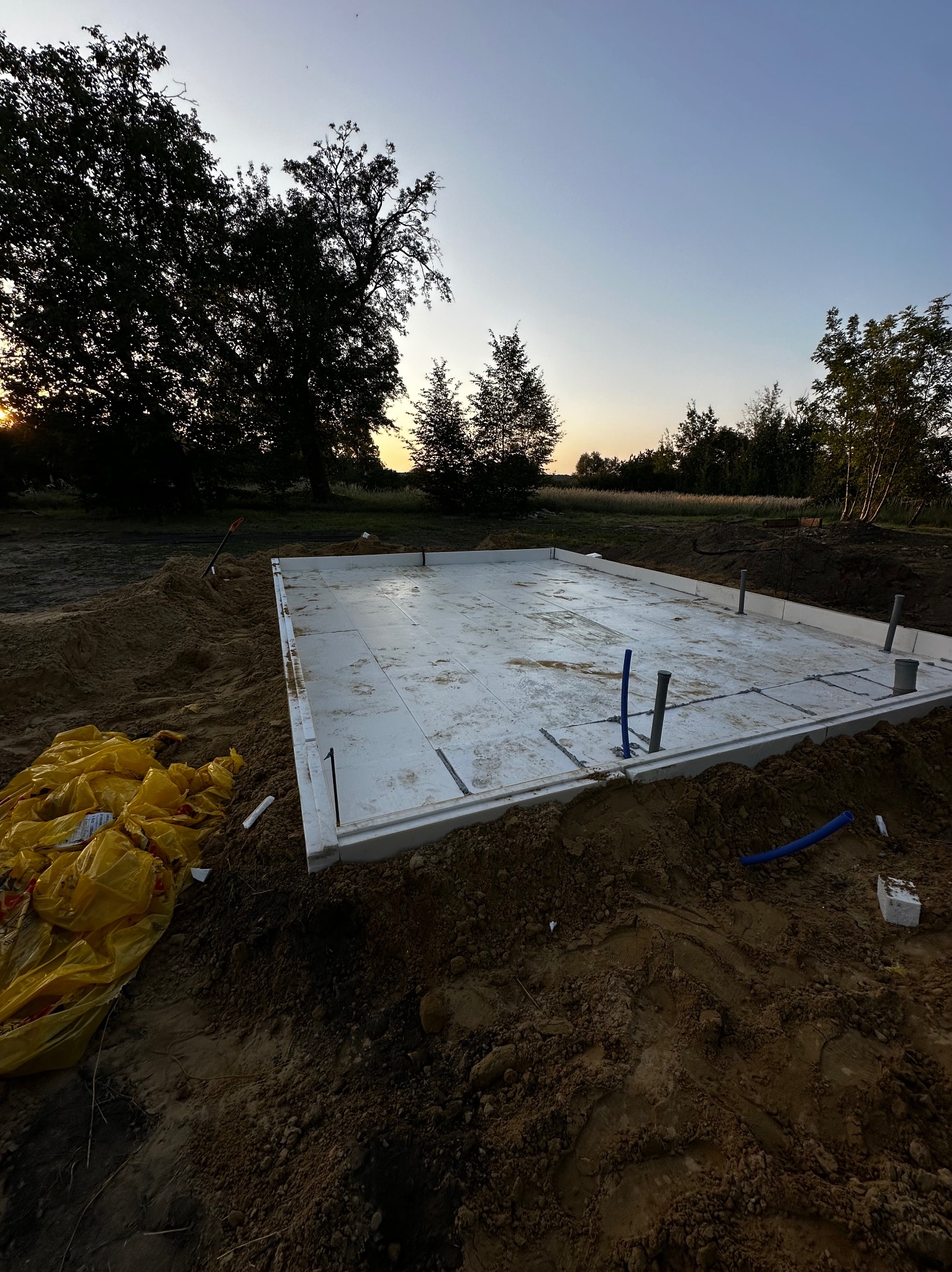 Przygotowany szalunek z płyt styropianowych pod płytę fundamentową na gruncie, z widocznymi pionowymi rurami instalacyjnymi i stertą żółtych worków z ziemią w tle, oświetlone zachodzącym słońcem.