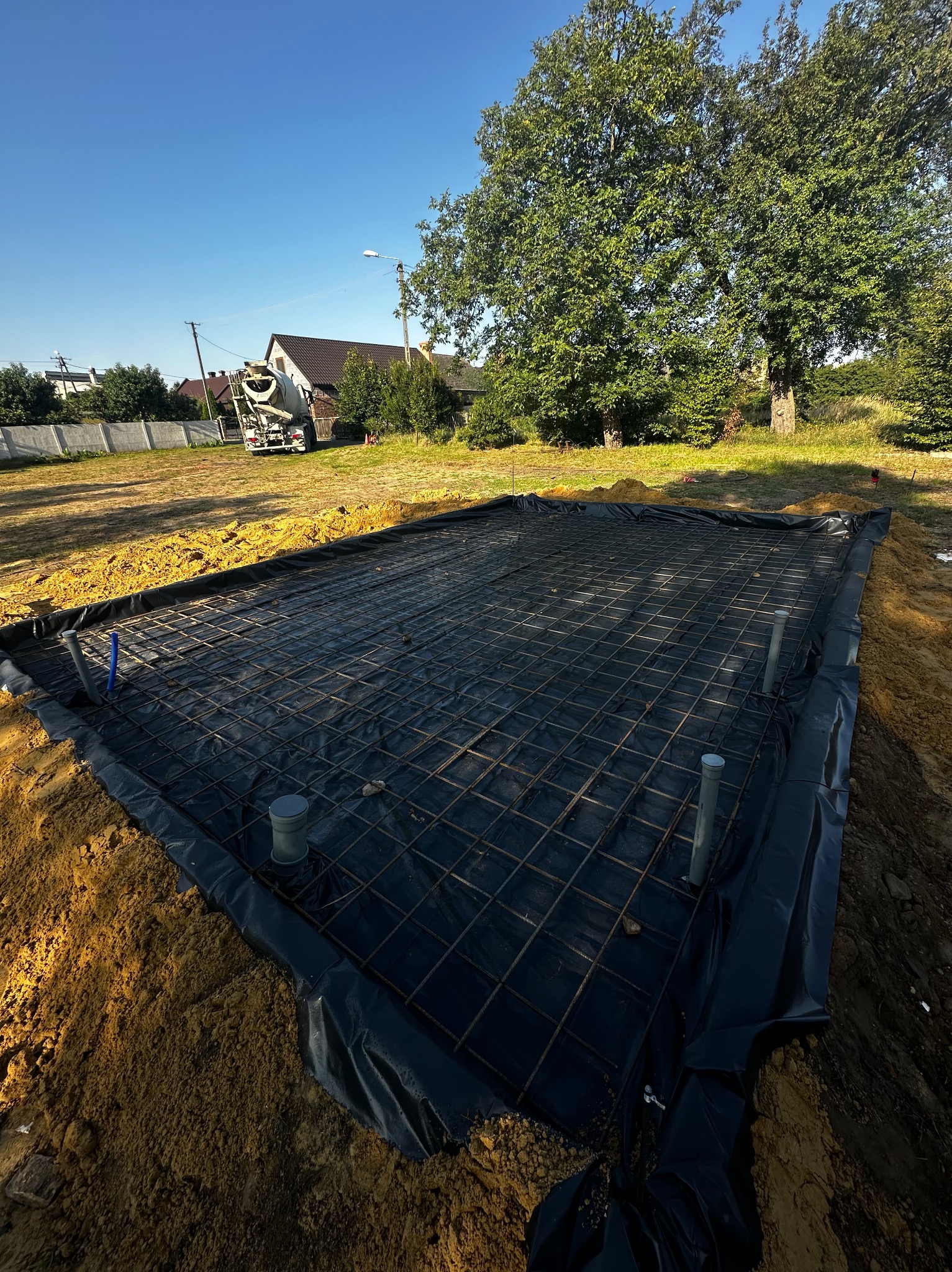 Zbrojenie przygotowane pod wylewkę fundamentów, widoczne czarne folie izolacyjne i rury kanalizacyjne, w tle betoniarka i domy jednorodzinne.