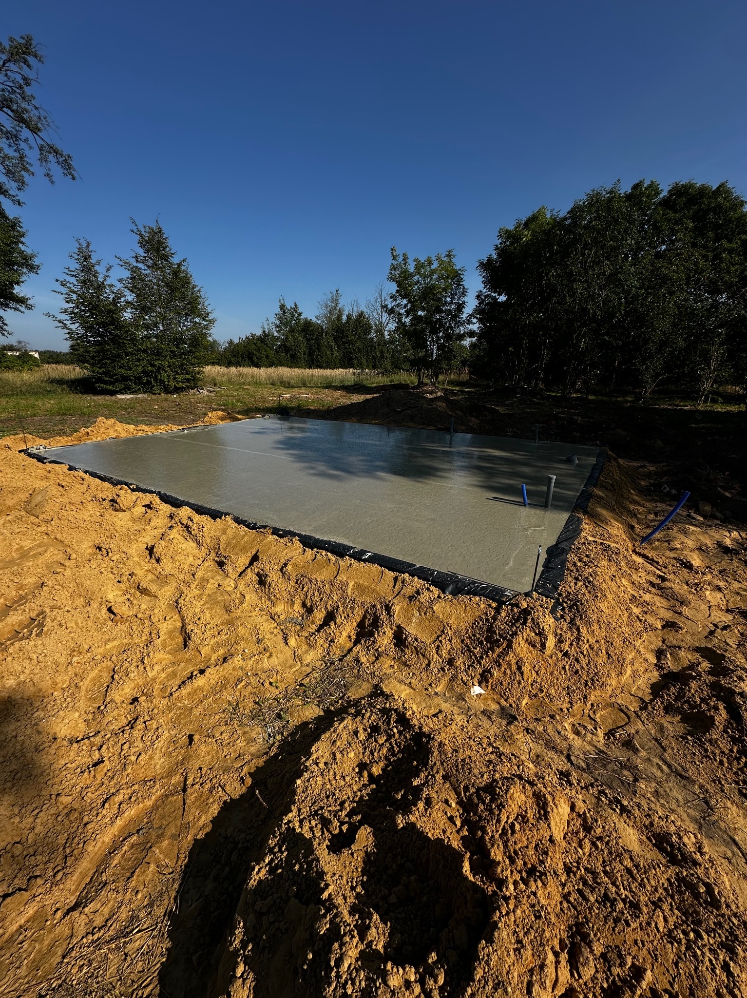 Wylany betonowy fundament pod budynek, widoczne wystające rury instalacyjne, otoczone wykopem z ziemi.