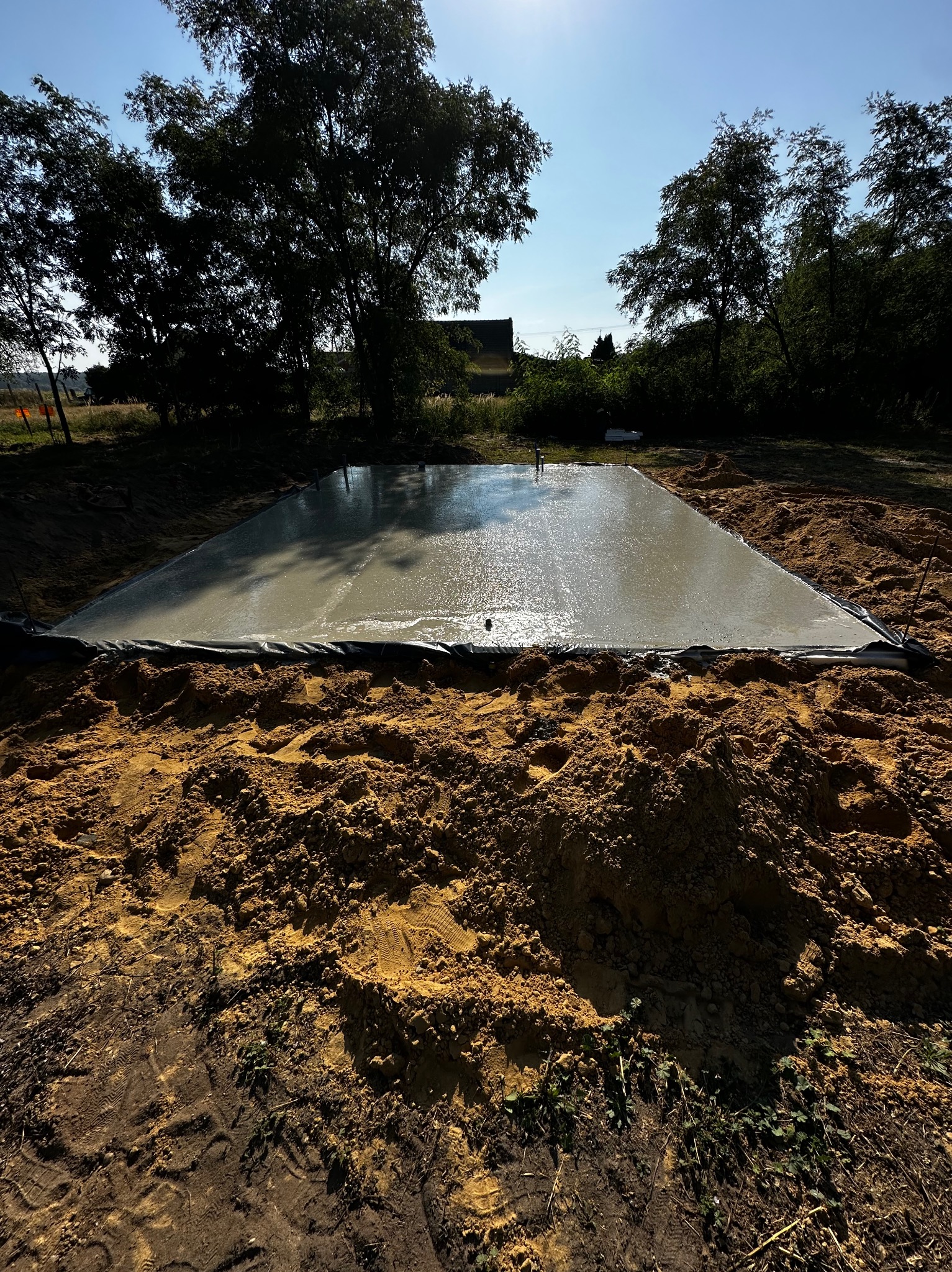Świeżo wylana, mokra płyta fundamentowa na czarnej folii, otoczona ziemią, w tle drzewa i błękitne niebo.