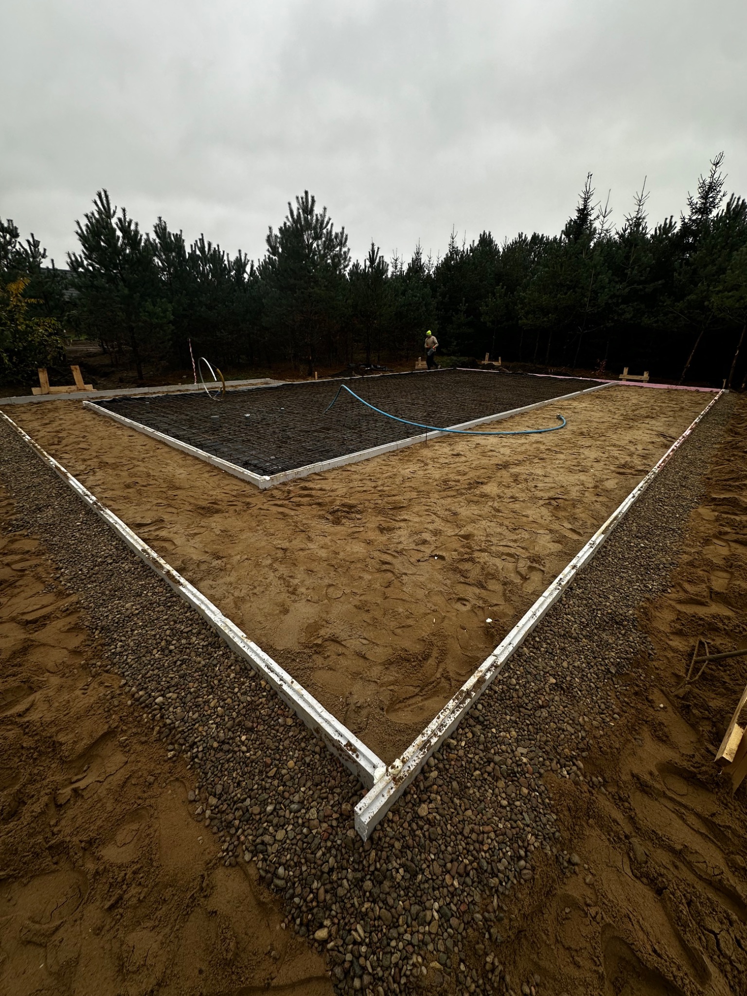 Przygotowany teren pod wylewkę fundamentów z widocznym zbrojeniem i obrysem szalunku, otoczony piaskiem i drobnym żwirem, w tle las iglasty i pochmurne niebo.