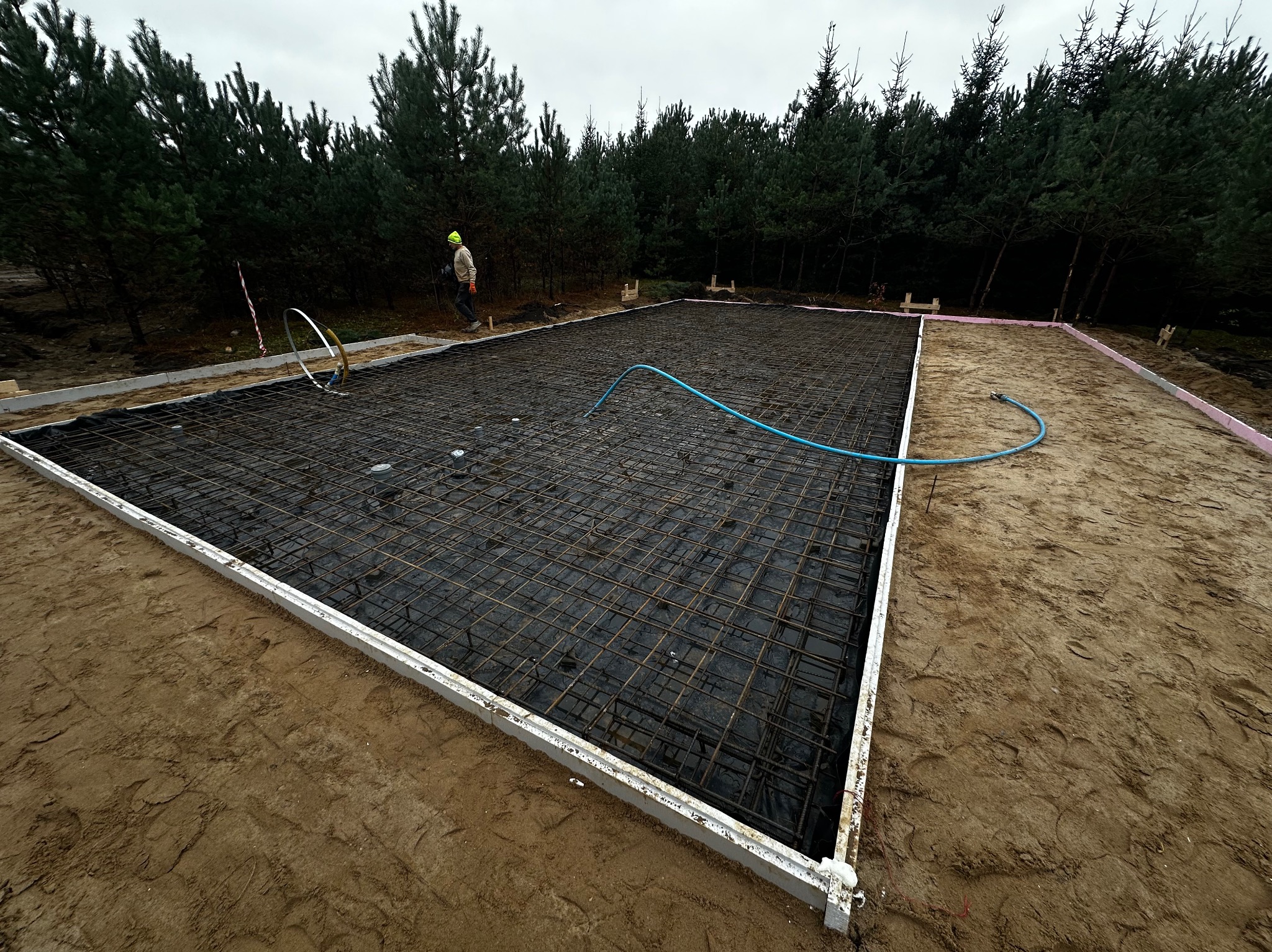 Przygotowany szalunek i zbrojenie pod płytę fundamentową na działce z widocznym pracownikiem w oddali.