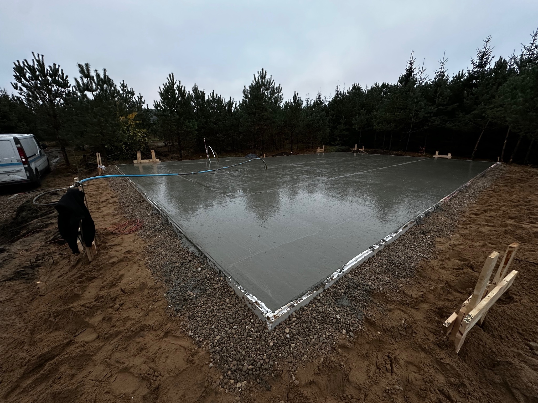 Świeżo wylana płyta fundamentowa na gruncie, widoczne szalunki i otaczający teren z drzewami w tle.