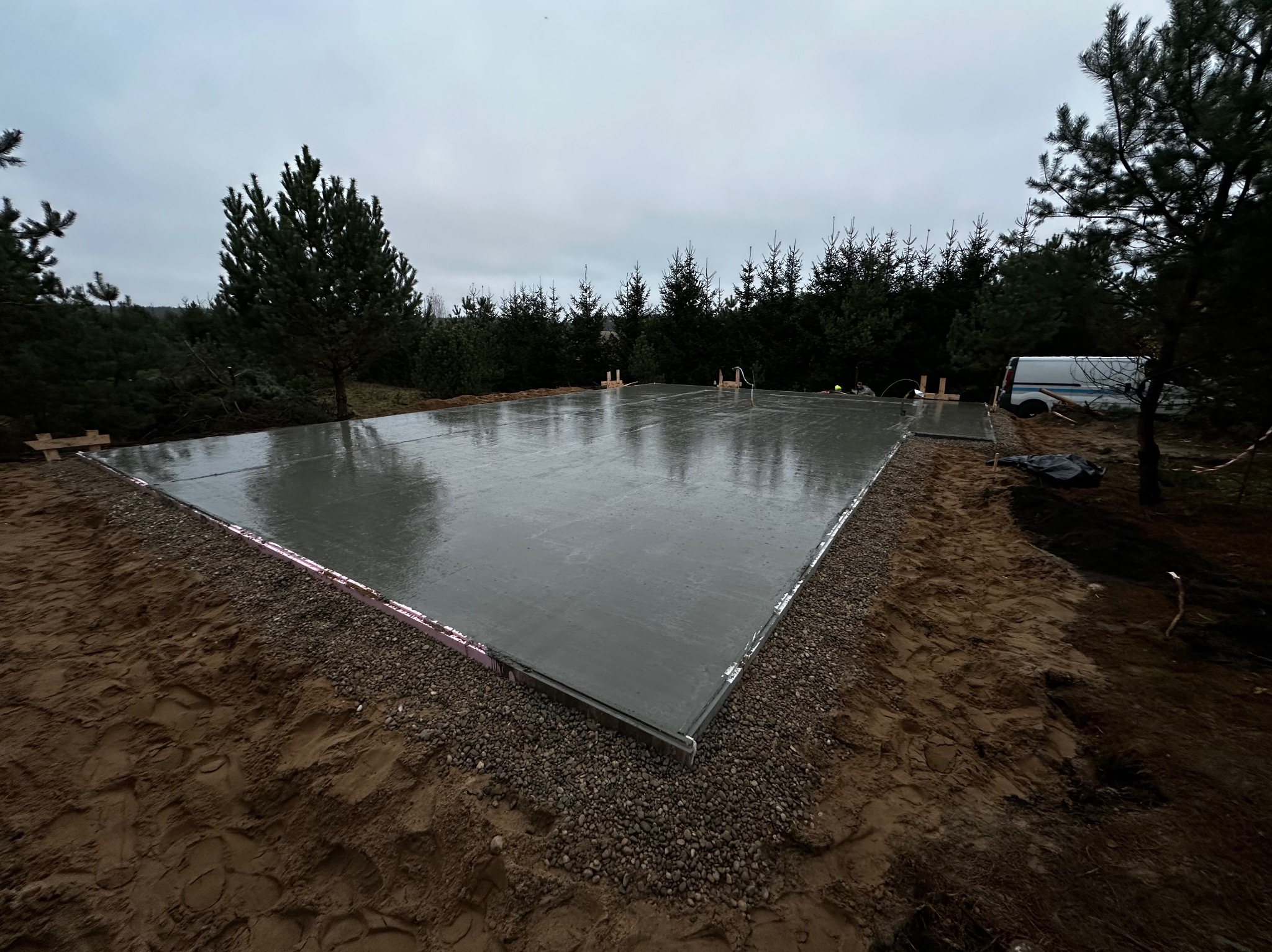 Świeżo wylana, gładka płyta fundamentowa z widoczną izolacją obwodową ze styropianu, otoczona ziemią i drobnym żwirem, na tle zalesionego terenu i pochmurnego nieba.