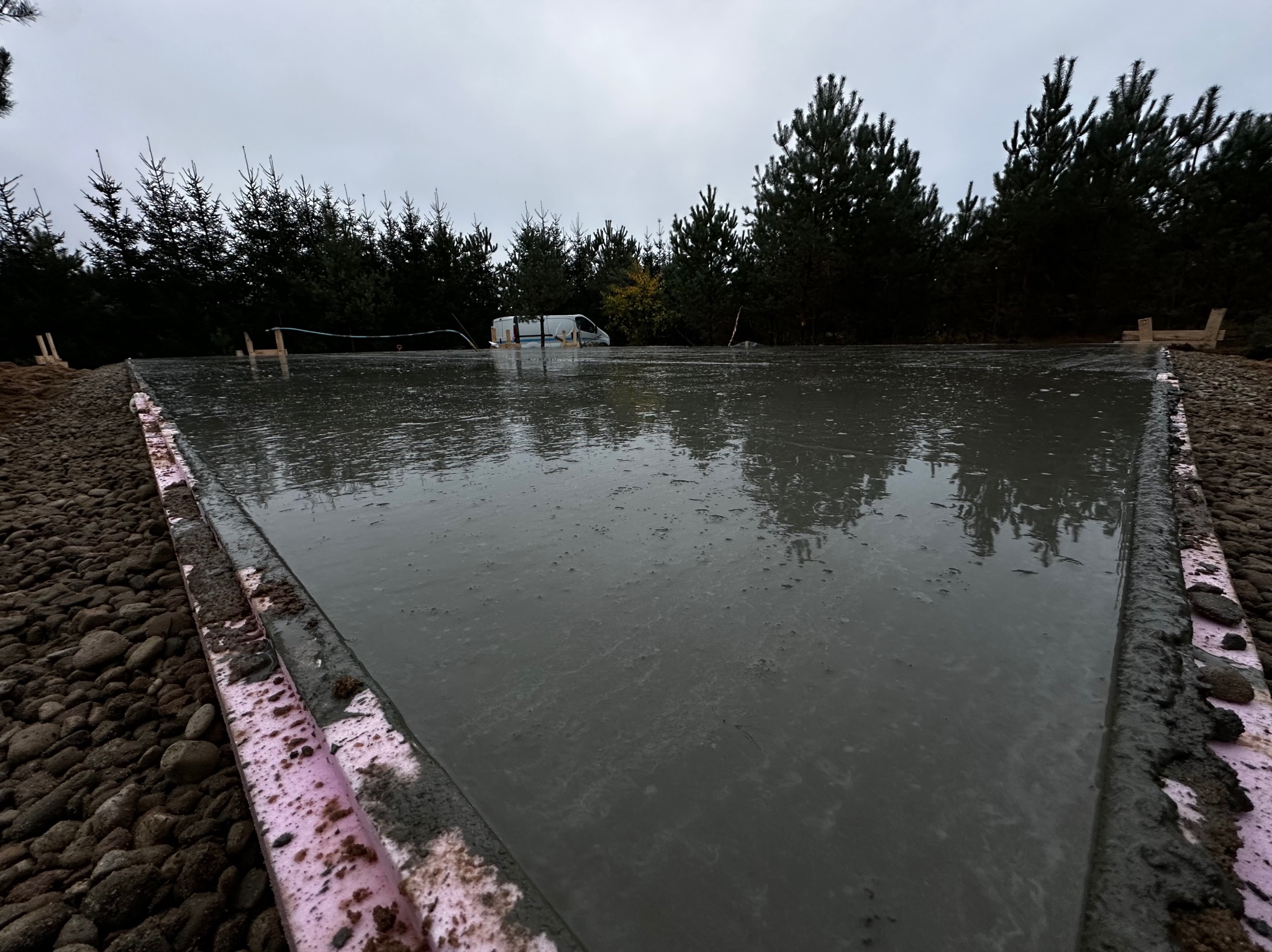 Świeżo wylany beton na przygotowanym fundamencie, widoczne szalunki z różowego styropianu i otaczające kamienie, w tle las iglasty i zaparkowany bus.
