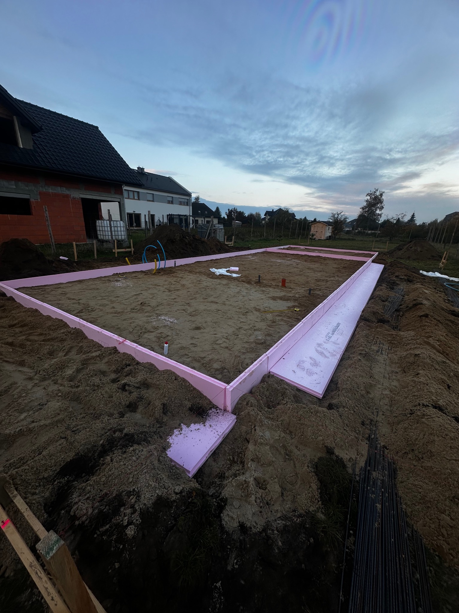 Przygotowany teren pod fundamenty domu jednorodzinnego z widocznym szalunkiem z różowego styropianu, rury instalacyjne i zbrojenie w tle, sfotografowane o zmierzchu.