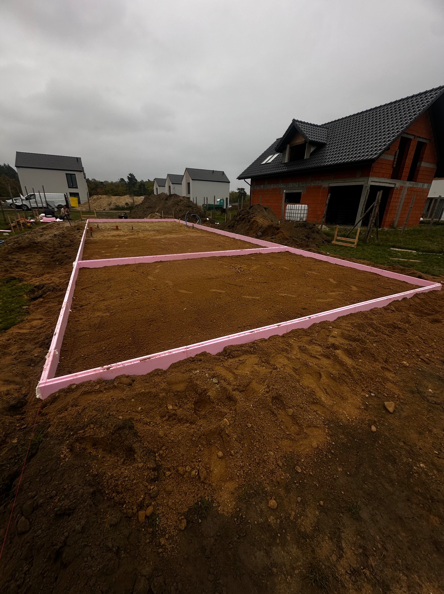 Przygotowany wykop z różową izolacją obwodową, widok na plac budowy z rozpoczętą budową domu z czerwonej cegły i okoliczną zabudową w tle.