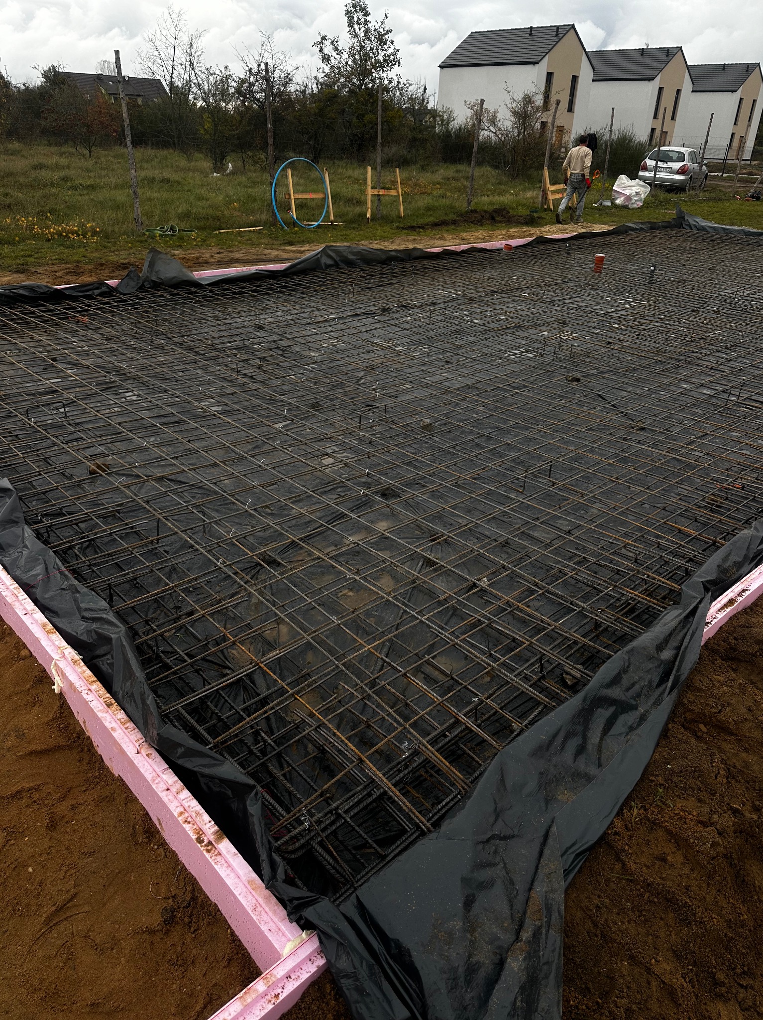Zbrojenie fundamentu z siatki stalowej na czarnej folii izolacyjnej, obłożone różowym styropianem, w tle budynki jednorodzinne i pracownik budowlany.