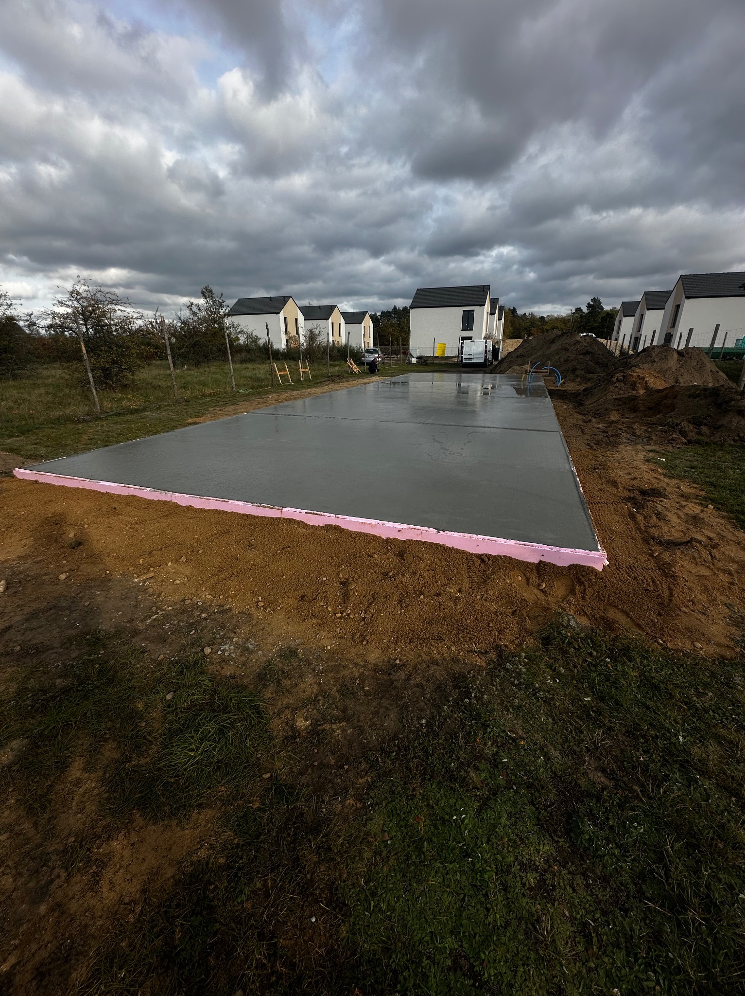 Wylany fundament z różową izolacją termiczną na obrzeżach, na tle szeregu nowoczesnych domów jednorodzinnych pod pochmurnym niebem.