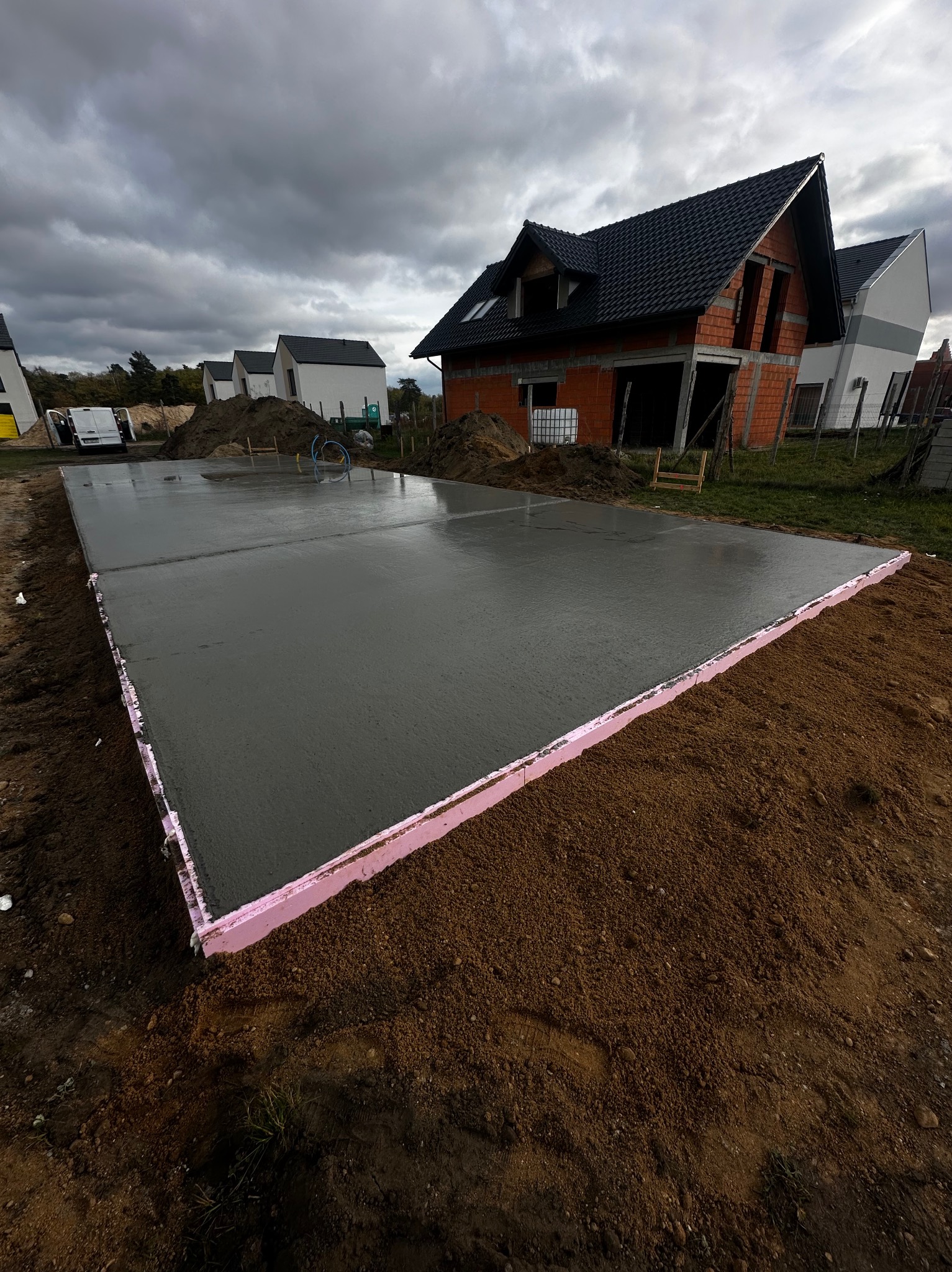 Świeżo wylana płyta fundamentowa z różowym styropianem obwodowym, w tle budynek w trakcie budowy z czerwonej cegły i szare, pochmurne niebo.