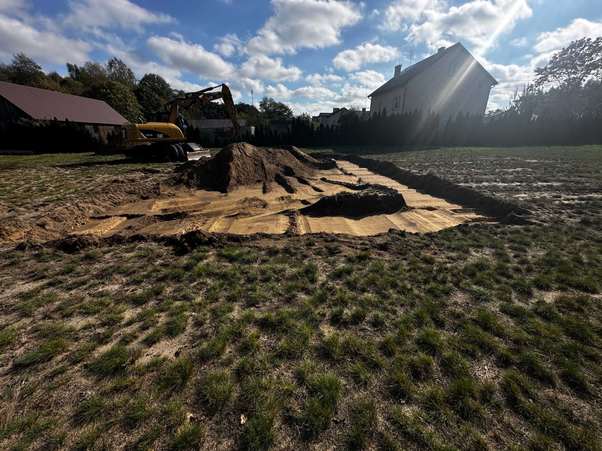 Przygotowany teren pod budowę z widocznym wykopem, wyrównanym podłożem i stertą ziemi w tle, obok stoi żółta koparka CAT, w oddali zabudowania.