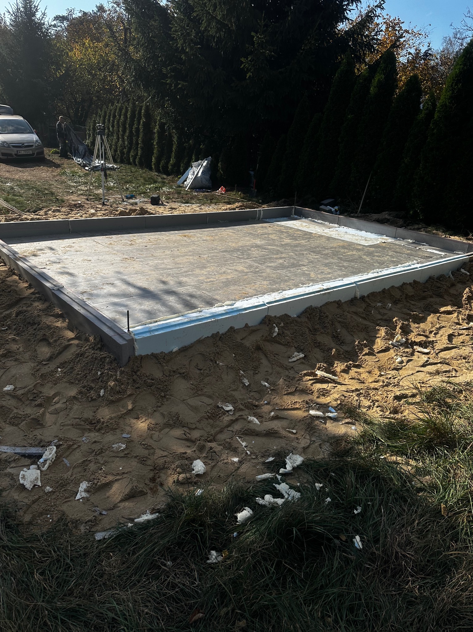 Przygotowany fundament pod budowę domu jednorodzinnego, widoczna płyta fundamentowa z szalunkiem i izolacją, w tle drzewa i częściowo widoczny samochód.