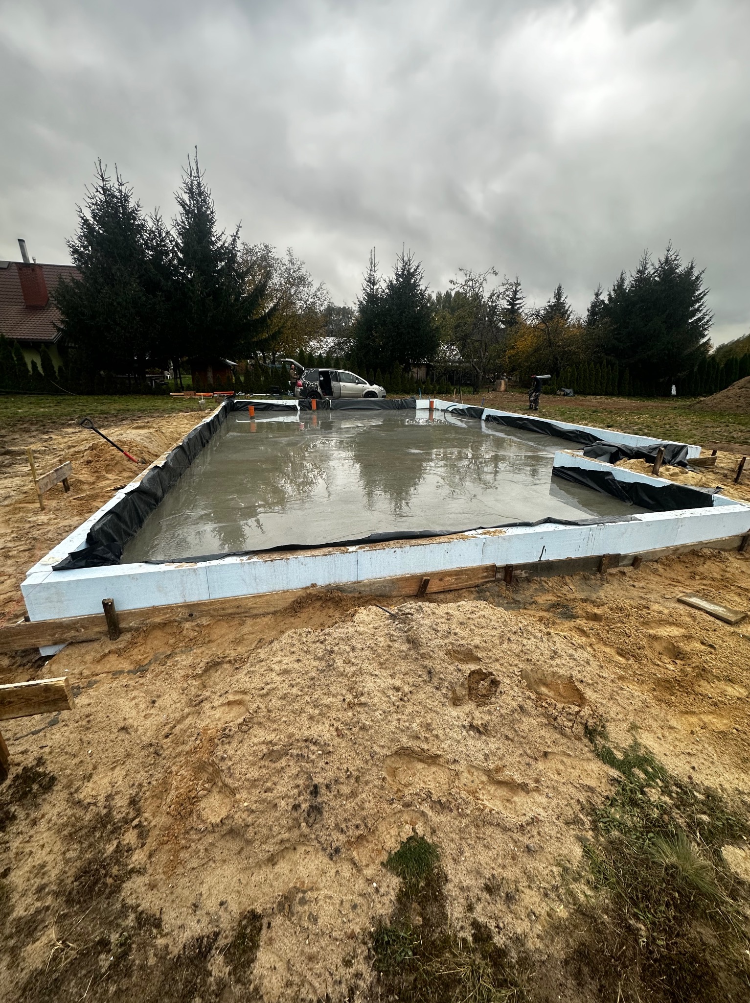 Wylany fundament z izolacją i szalunkiem styropianowym na piaszczystym gruncie, widoczne drzewa w tle i pochmurne niebo.