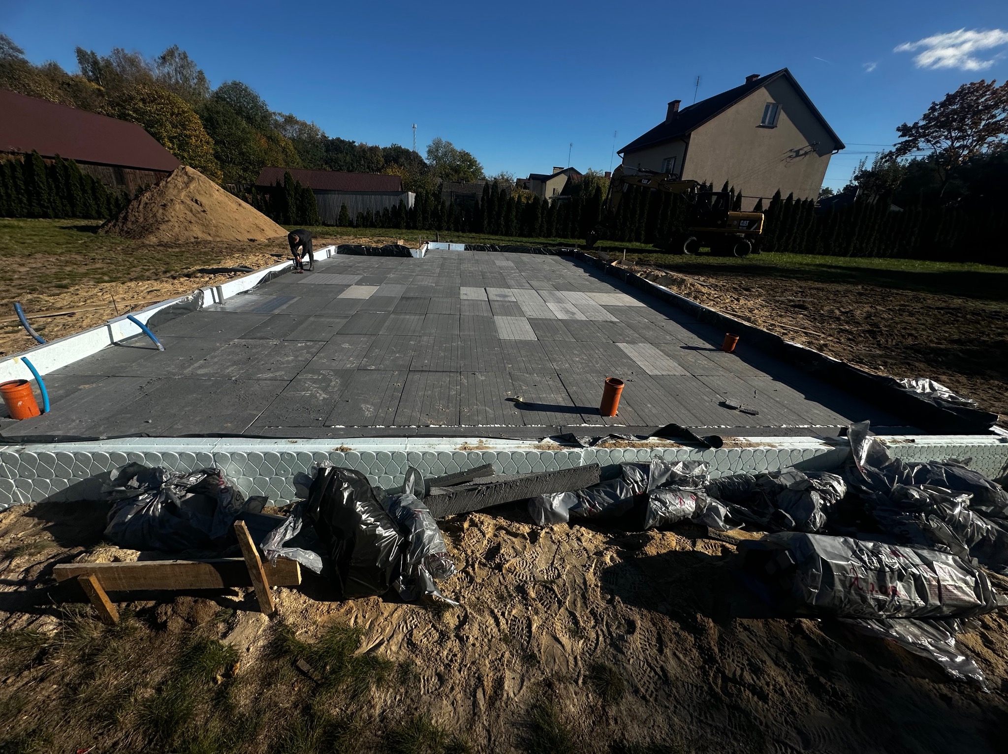 Przygotowany teren pod fundamenty z ułożoną izolacją termiczną i folią, widoczny wykop z workami i resztkami materiałów budowlanych, w tle budynek mieszkalny i koparka.