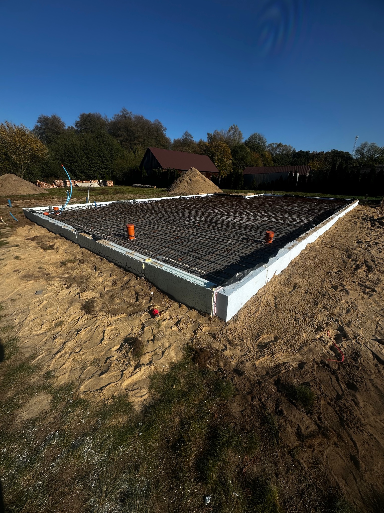 Przygotowany teren pod budowę domu: wylany fundament z izolacją, zbrojony stalową siatką, widoczne rury instalacyjne, w tle pryzma piasku i budynek gospodarczy.