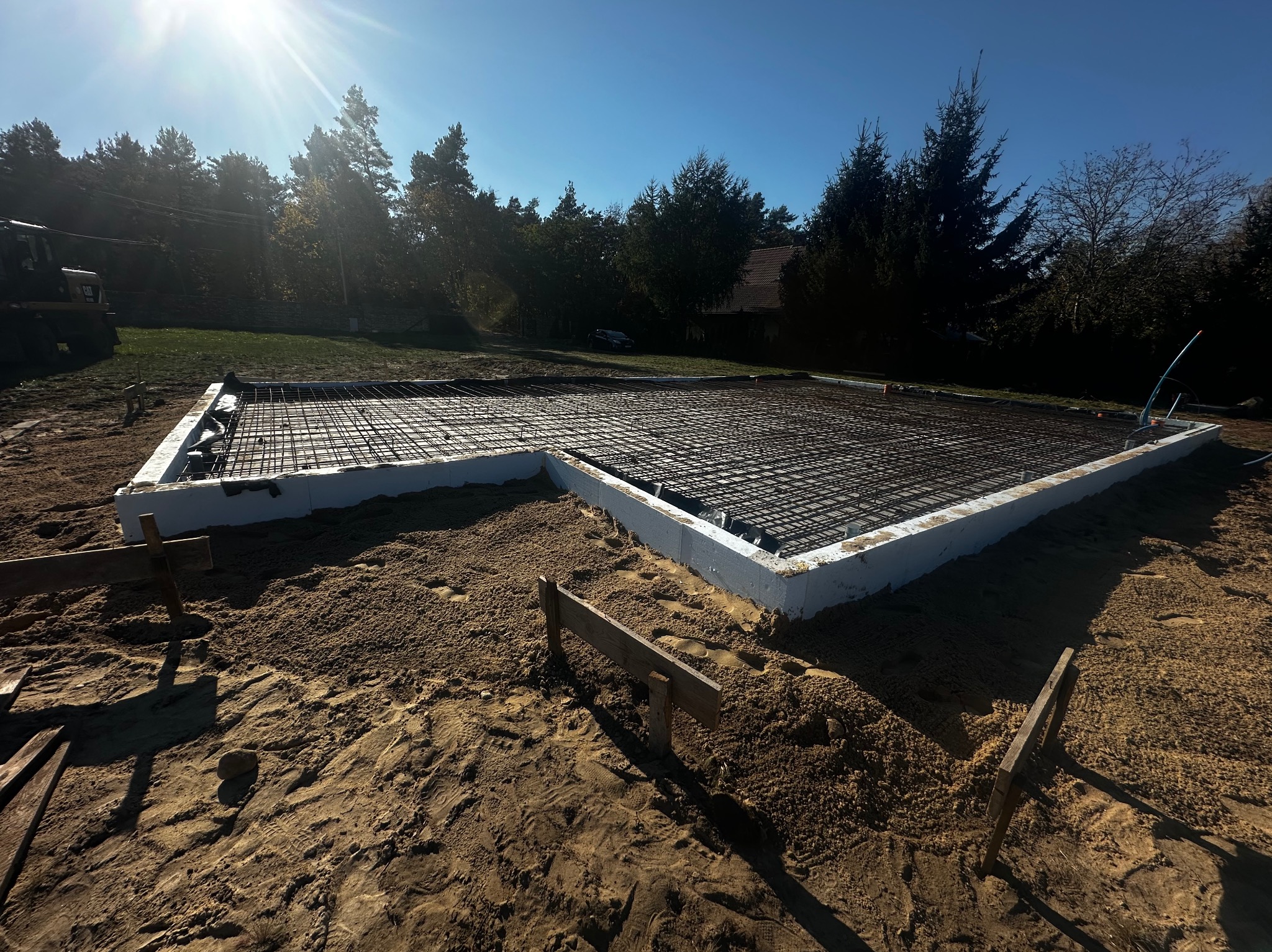 Przygotowany zbrojony szalunek pod płytę fundamentową na piaszczystym podłożu, widoczne drewniane elementy stabilizujące, w tle drzewa i błękitne niebo.