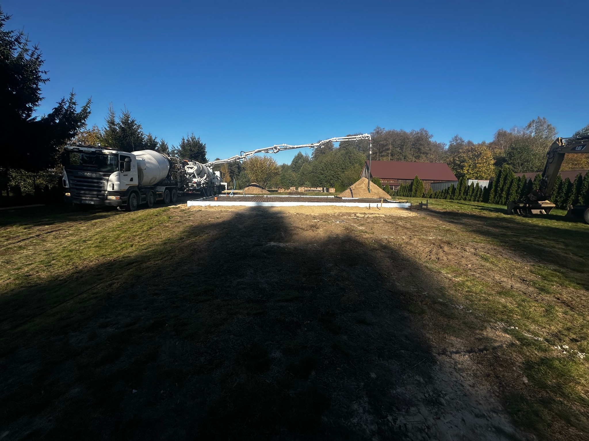 Wylewanie betonu na przygotowany zbrojony fundament pod budynek, z użyciem pompy do betonu i betonomieszarki, w tle zielona trawa i drzewa.