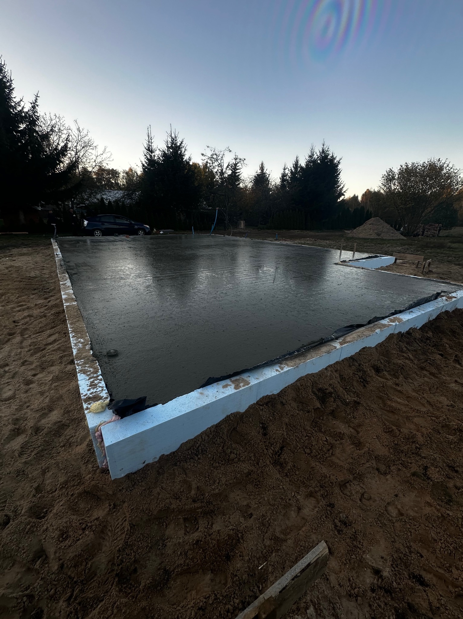 Wylana płyta fundamentowa z izolacją termiczną, widok z góry, w tle drzewa i samochód.