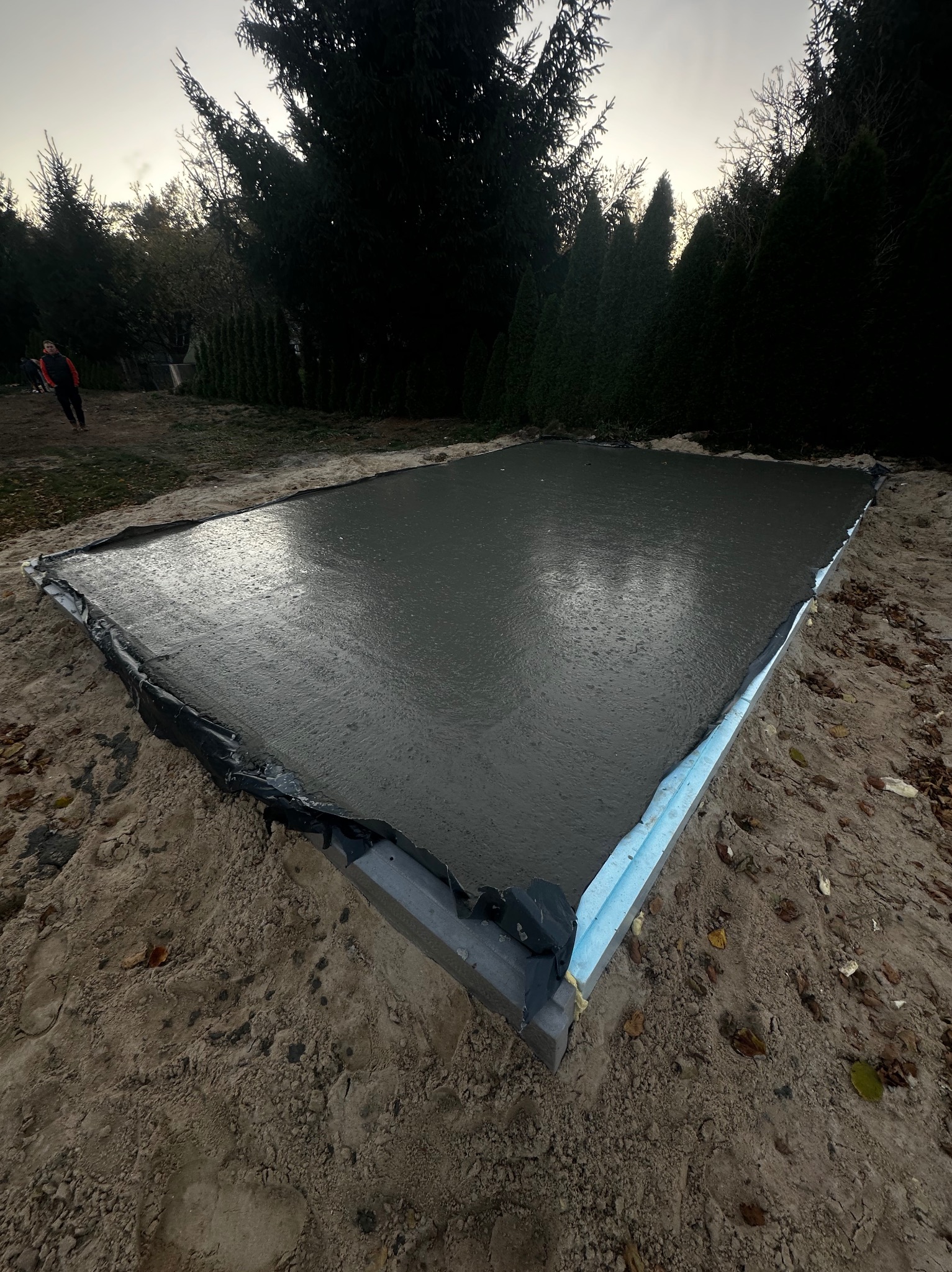 Świeżo wylany beton na przygotowanym gruncie pod fundamenty, widoczne szalunki z izolacją, osoba w oddali.