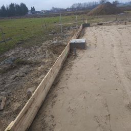 PPSW - Przygotowanie terenu pod fundament: drewniane szalunki ustawione wzdłuż wykopu, widoczne betonowe elementy i rury, w tle sterta ziemi i krajobraz wiejski w pochmurny dzień.