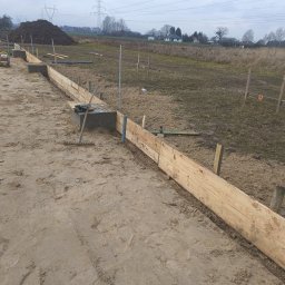 PPSW - Przygotowanie szalunku pod fundament z widocznymi drewnianymi deskami, betonowymi bloczkami, poziomica i zagęszczarką gruntu na piaszczystym podłożu, w tle pole i słupy wysokiego napięcia.