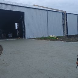 PPSW - Świeżo wylana, gładka, betonowa nawierzchnia przed halą przemysłową z otwartymi wrotami i materiałami budowlanymi w środku.