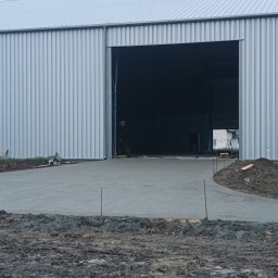 PPSW - Świeżo wylana, szara posadzka przed budynkiem przemysłowym z blachy trapezowej, widoczny pracownik wewnątrz budynku, wystające pręty zbrojeniowe w ziemi.