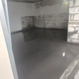 Świeżo wylana, gładka, szara posadzka w surowym pomieszczeniu z niedokończonymi ścianami z betonu komórkowego i częściowo otynkowanymi fragmentami, widoczne gniazdka elektryczne i lampa sufitowa.