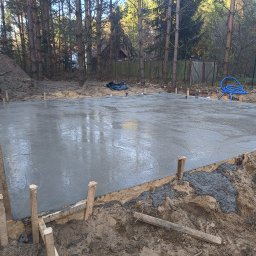 PPSW - Świeżo wylana, gładka płyta fundamentowa z drewnianym szalunkiem i wystającymi kołkami, otoczona ziemią i drzewami w tle.