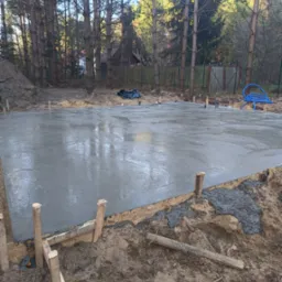 Świeżo wylana, gładka płyta fundamentowa z drewnianym szalunkiem i wystającymi kołkami, otoczona ziemią i drzewami w tle.