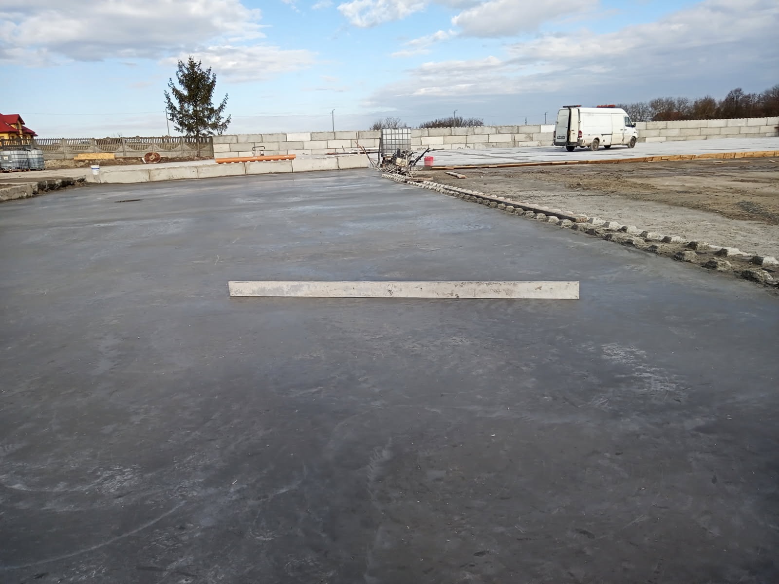 Świeżo wylana, gładka, szara posadzka z widoczną łatą poziomującą, w tle budynek, drzewo i biały samochód dostawczy.