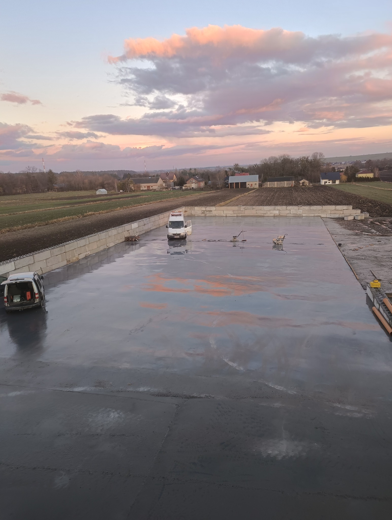 Świeżo wylana, gładka posadzka betonowa na zewnątrz, z widocznymi śladami po maszynach do wygładzania, w tle dwa samochody dostawcze i zachodzące słońce odbijające się w mokrej powierzchni.