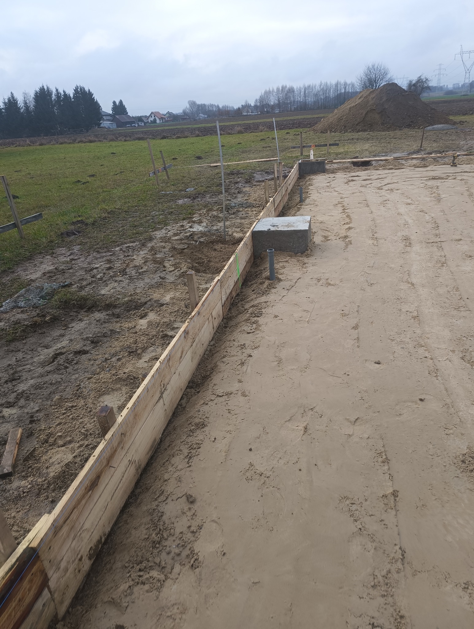 Przygotowanie terenu pod fundament: drewniane szalunki ustawione wzdłuż wykopu, widoczne betonowe elementy i rury, w tle sterta ziemi i krajobraz wiejski w pochmurny dzień.