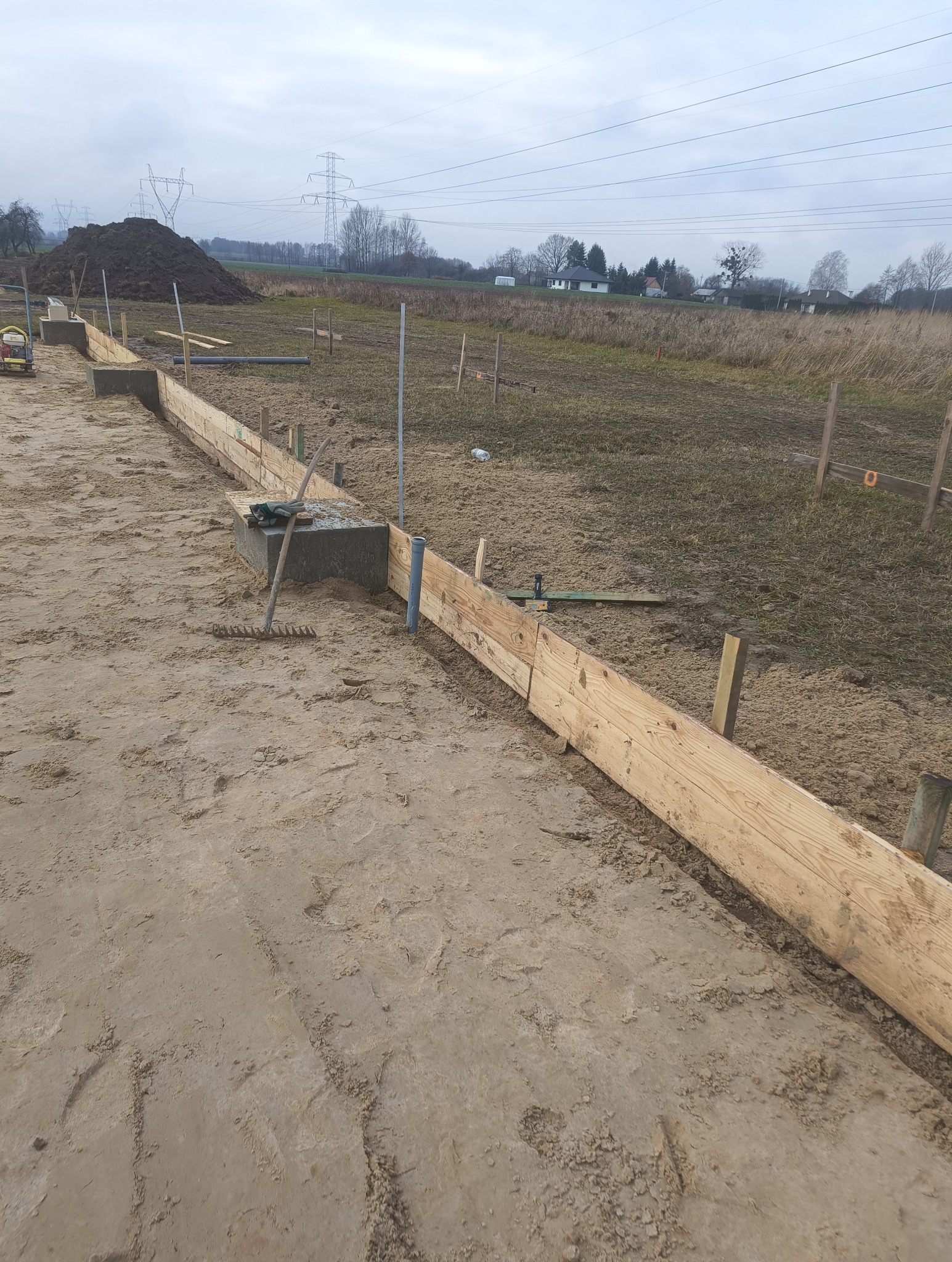 Przygotowanie szalunku pod fundament z widocznymi drewnianymi deskami, betonowymi bloczkami, poziomica i zagęszczarką gruntu na piaszczystym podłożu, w tle pole i słupy wysokiego napięcia.