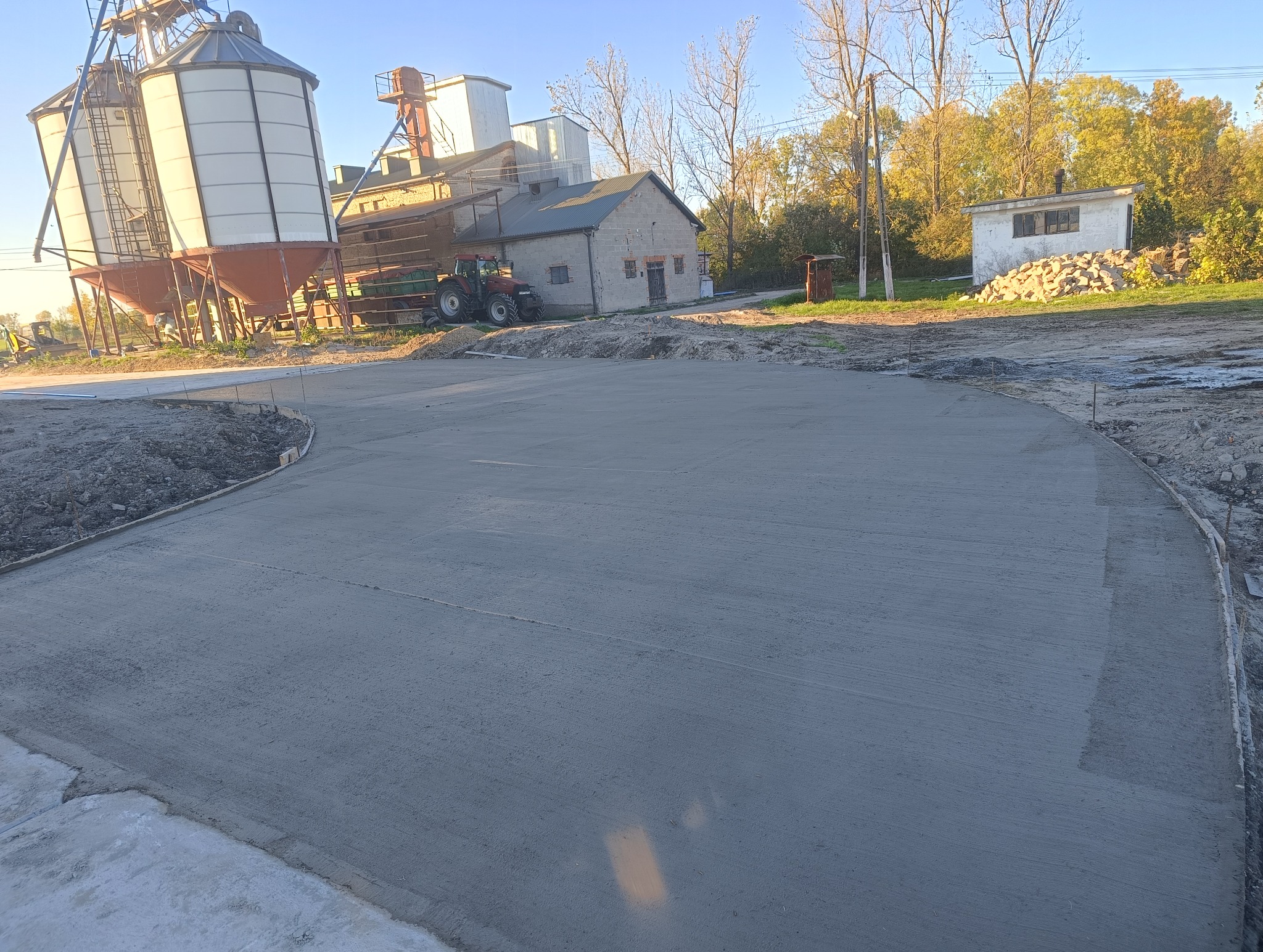 Świeżo wylana, gładka betonowa nawierzchnia placu manewrowego w gospodarstwie rolnym, z widocznymi szalunkami i zbrojeniem na obrzeżach, w tle budynki gospodarcze i silosy.