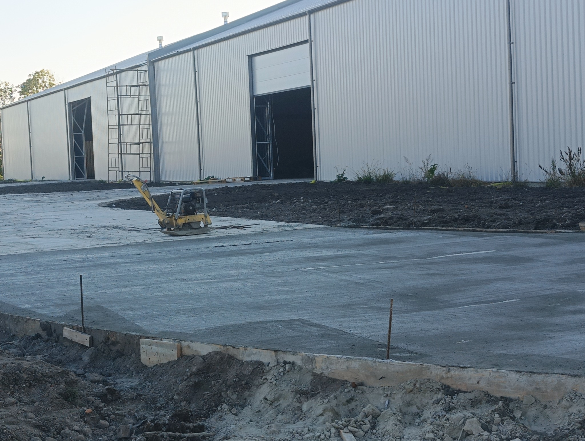 Świeżo wylana, szara betonowa posadzka przemysłowa przed budynkiem magazynowym z blachy trapezowej. W tle widoczne rusztowanie i otwarte wrota magazynu. Na posadzce stoi ubijarka wibracyjna.