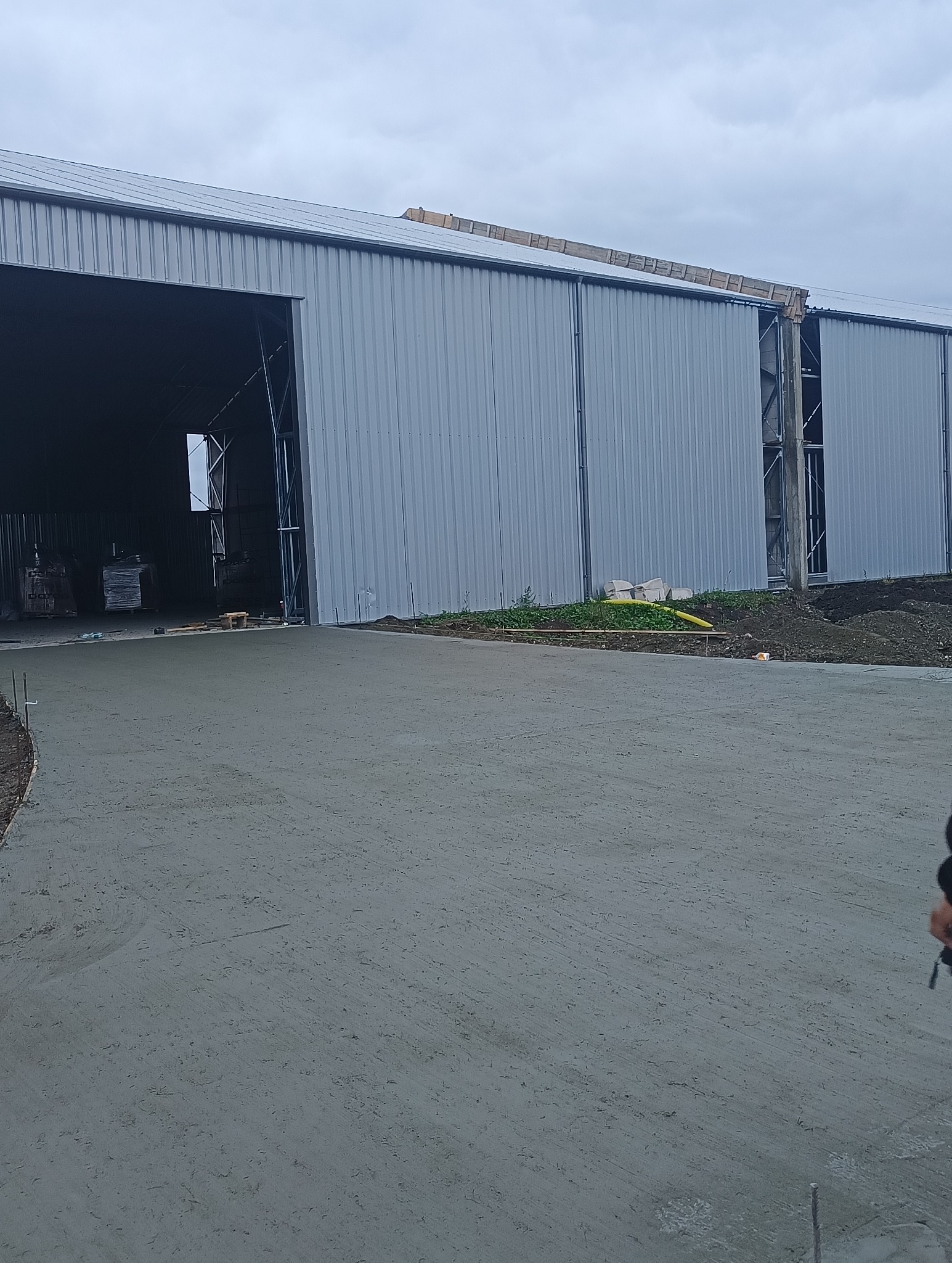 Świeżo wylana, gładka, betonowa nawierzchnia przed halą przemysłową z otwartymi wrotami i materiałami budowlanymi w środku.