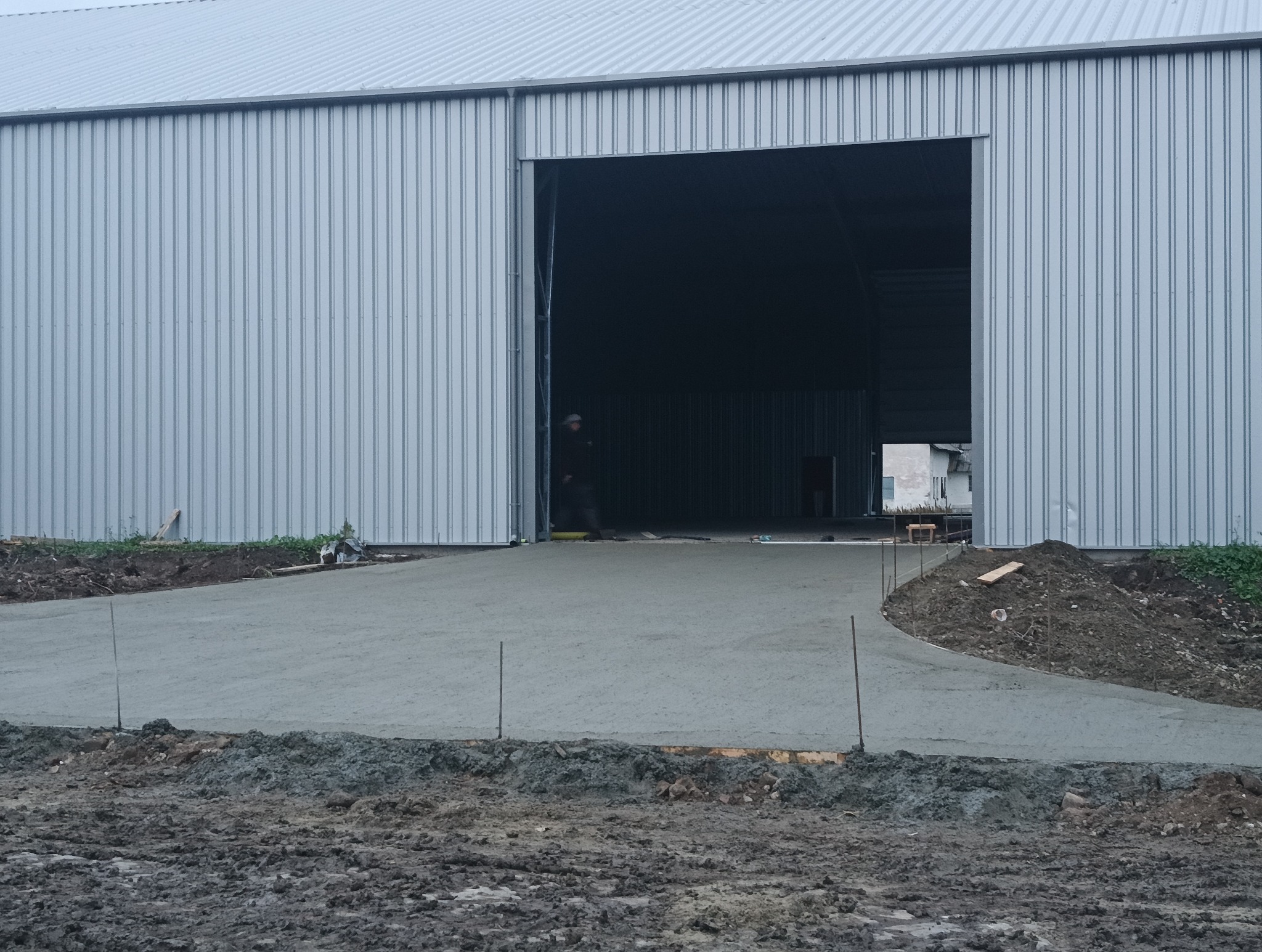 Świeżo wylana, szara posadzka przed budynkiem przemysłowym z blachy trapezowej, widoczny pracownik wewnątrz budynku, wystające pręty zbrojeniowe w ziemi.