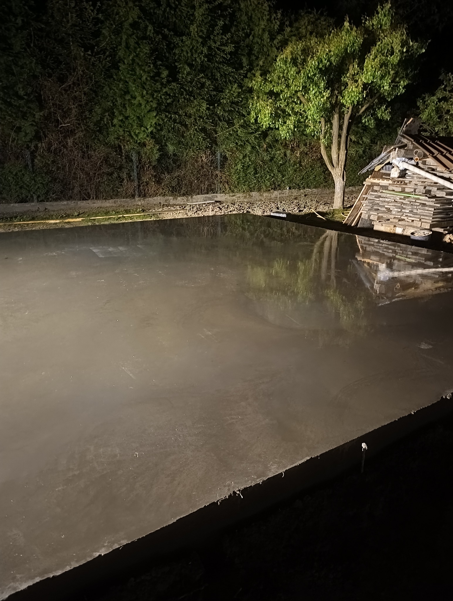 Świeżo wylana, mokra betonowa płyta fundamentowa w ciemnym otoczeniu, odbijająca światło, z widocznymi deskami i stertą materiałów budowlanych w tle.